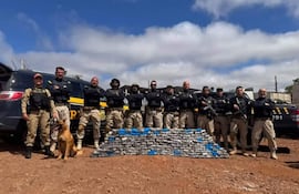 Mujer en uniforme táctico frente a agentes en uniforme negro, con un perro. Paquetes de droga detrás en un terreno arenoso.