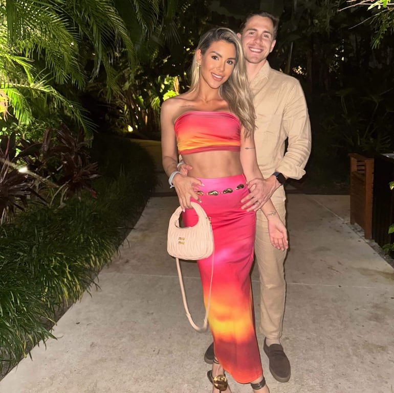 Mujer sonriente con vestido colorido y bolso Miu Miu, acompañada por un hombre con atuendo claro, en un entorno tropical.