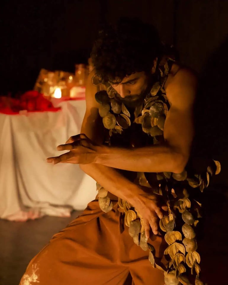 Hombre de cabello rizado y barba, vestido con pantalones marrones, actuando en un escenario con iluminación tenue y decoraciones.