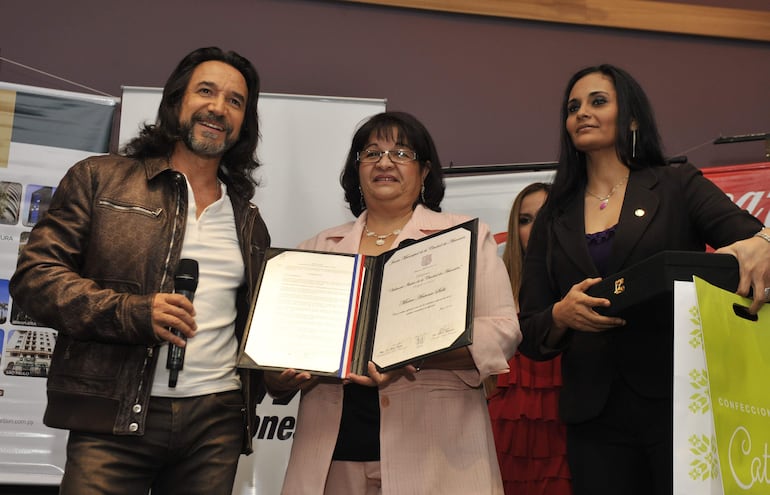 Marco Antonio Solís sonríe con chaqueta marrón, mientras dos mujeres lo acompañan sosteniendo documentos y una caja en un ambiente formal.