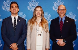 Mujer en vestido blanco y chaqueta beige flanqueada por hombres en trajes oscuros, todos sonriendo en un evento formal.