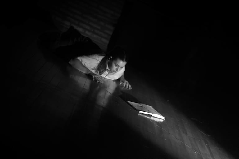 Mujer inclinada hacia un cuaderno iluminado, vestida con camiseta clara y cabello recogido, en ambiente oscuro y de reflexión.