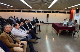 Cuatro hombres sentados en una mesa discuten mientras una audiencia los escucha atentamente en un salón bien iluminado.
