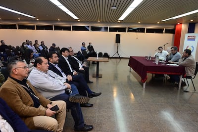 Cuatro hombres sentados en una mesa discuten mientras una audiencia los escucha atentamente en un salón bien iluminado.