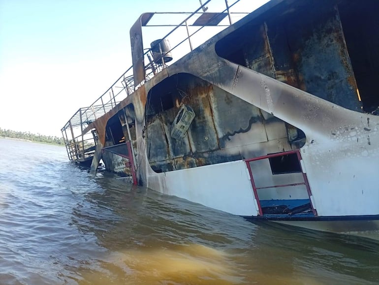 Barco semi sumergido y dañado por fuego, con costados quemados y ambiente natural de árboles al fondo.