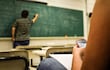 Estudiante con teléfono móvil en aula, sentado al lado de una libreta, mientras el docente escribe en la pizarra.