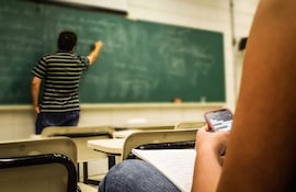 Estudiante con teléfono móvil en aula, sentado al lado de una libreta, mientras el docente escribe en la pizarra.