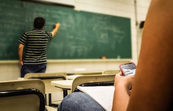 Estudiante con teléfono móvil en aula, sentado al lado de una libreta, mientras el docente escribe en la pizarra.