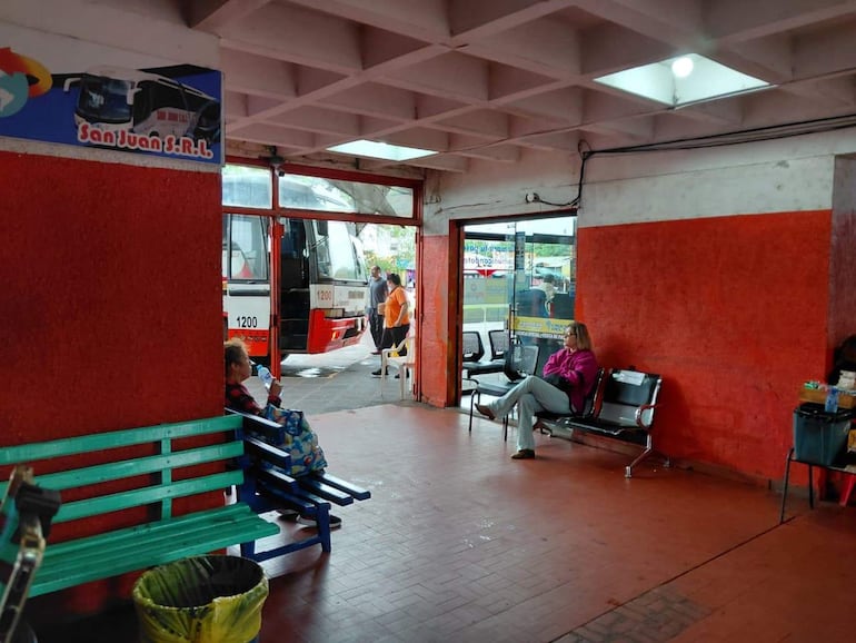 Mujer mayor sentada con helado en banco verde, grupo de personas esperando bus en terminal deteriorada.