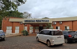 Fachada del hospital con letrero 'URGENCIAS PEDIÁTRICAS', vehículos estacionados y clima nublado.