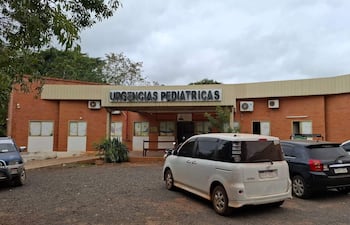 Fachada del hospital con letrero 'URGENCIAS PEDIÁTRICAS', vehículos estacionados y clima nublado.