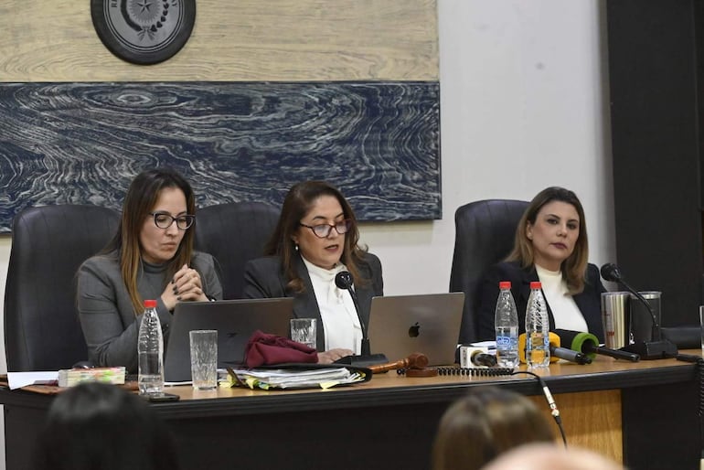 Tres mujeres sentadas, una en abrigo oscuro y blusa blanca hablando, mientras las otras escuchan atentamente en ambiente formal.