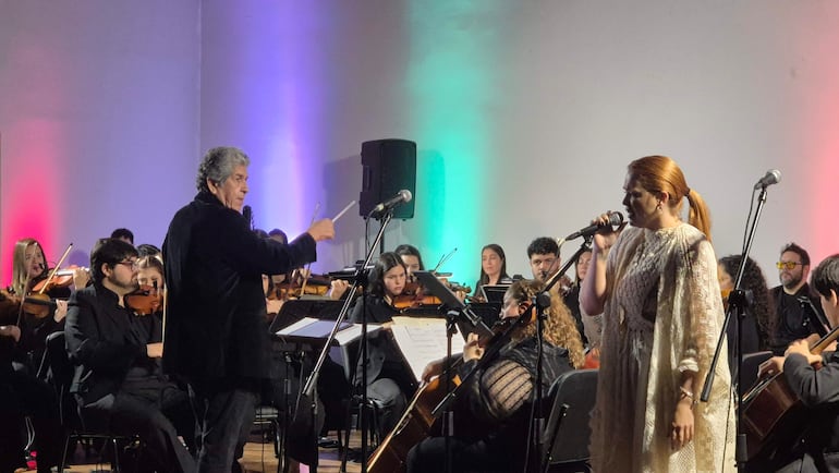 Directores de orquesta y músicos en escena. Una cantante en vestido claro sostiene un micrófono, iluminados con luces verdes y rojas.