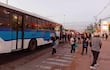 Personas en una estación de buses, algunos con mochilas, esperando el transporte. Autobús azul y blanco visible en el fondo.