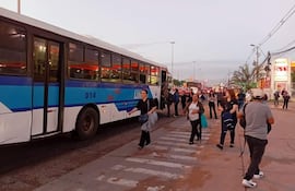 Personas en una estación de buses, algunos con mochilas, esperando el transporte. Autobús azul y blanco visible en el fondo.