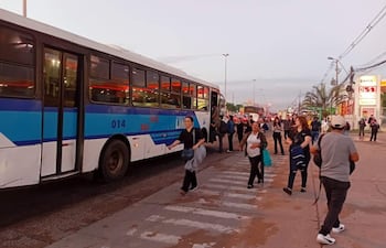 Personas en una estación de buses, algunos con mochilas, esperando el transporte. Autobús azul y blanco visible en el fondo.
