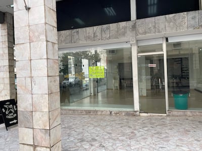 Local comercial vacío con sillas apiladas, cartel de alquiler en la entrada y ambiente desolado.