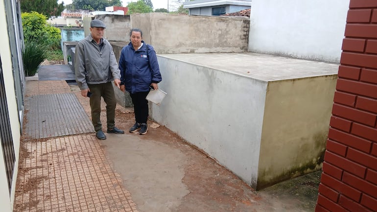 Felicia Ramona Benítez Aguilera en chaqueta azul, interactúa con un hombre de abrigo gris en un pasillo al aire libre.