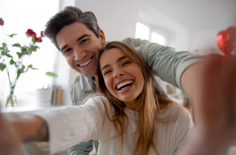 Hombre con cabello corto y castaño en camiseta clara y chaqueta, mujer con cabello largo y castaño en blusa blanca, ambos riéndose.