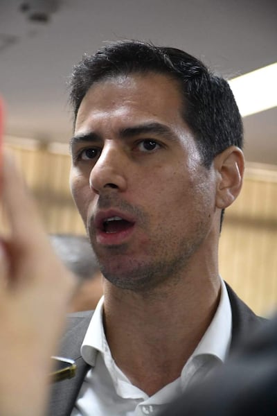 Alvaro Grau con camisa blanca y saco negro, gesticulando enérgicamente mientras otros manifiestan en el fondo.
