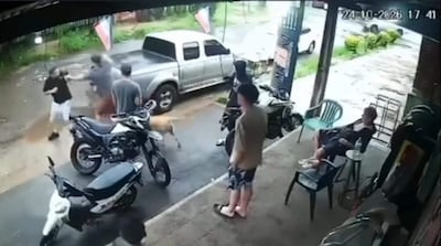 Un hombre de camiseta negra enfrenta a otro con camiseta gris en un entorno urbano, mientras motocicletas y un perro están cerca.