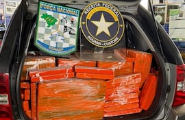 Maletero de vehículo lleno de paquetes de marihuana, con escudos de la Força Nacional y Receita Federal en el fondo.