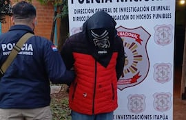Hombre con capucha roja es guiado por un policía con chaqueta estampada, en un ambiente exterior junto a un cartel de la Policía Nacional.