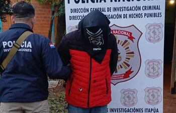 Hombre con capucha roja es guiado por un policía con chaqueta estampada, en un ambiente exterior junto a un cartel de la Policía Nacional.