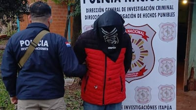 Hombre con capucha roja es guiado por un policía con chaqueta estampada, en un ambiente exterior junto a un cartel de la Policía Nacional.