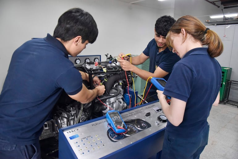 Dos hombres y una mujer en camisetas azul marino, ajustando motor y revisando diagnóstico en un taller mecánico.