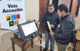 Hombre en chaqueta negra junto a máquina de votación, observando mientras sostiene un documento, ambiente electoral visible.