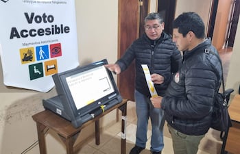 Hombre en chaqueta negra junto a máquina de votación, observando mientras sostiene un documento, ambiente electoral visible.