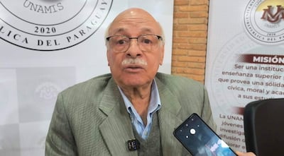 Pedro González, mayor y con gafas, en saco claro y camisa azul, sostiene un teléfono mientras habla ante un logo institucional.