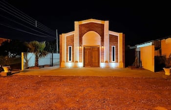 Edificación moderna con fachada de ladrillo iluminada por luces nocturnas, rodeada de terreno de tierra y una palmera a la izquierda.