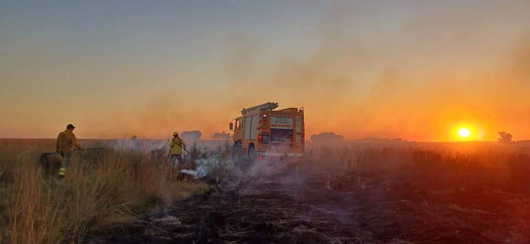 Tres bomberos, uno en uniforme marrón y otro en amarillo, luchan contra un incendio, con un camión de bomberos amarillo al fondo.