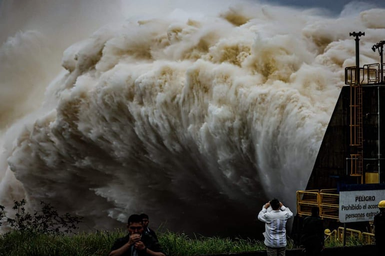 Hombres observan el agua marrón que desborda de la represa Itaipú, con señales de peligro alrededor.