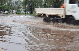 Camioneta blanca en movimiento sobre calle inundada en área urbana con vegetación alrededor.
