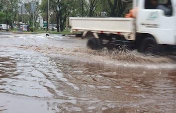 Camioneta blanca en movimiento sobre calle inundada en área urbana con vegetación alrededor.