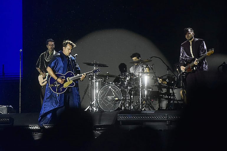 Cuatro músicos en el escenario: guitarrista eléctrico, vocalista con capa azul, y un baterista. Iluminación brillante y ambiente animado.