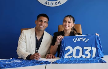 Diego Gómez con traje claro firma en una oficina, mientras una mujer sonríe sosteniendo camiseta azul con 'GOMEZ 2031'.