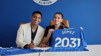 Diego Gómez con traje claro firma en una oficina, mientras una mujer sonríe sosteniendo camiseta azul con 'GOMEZ 2031'.