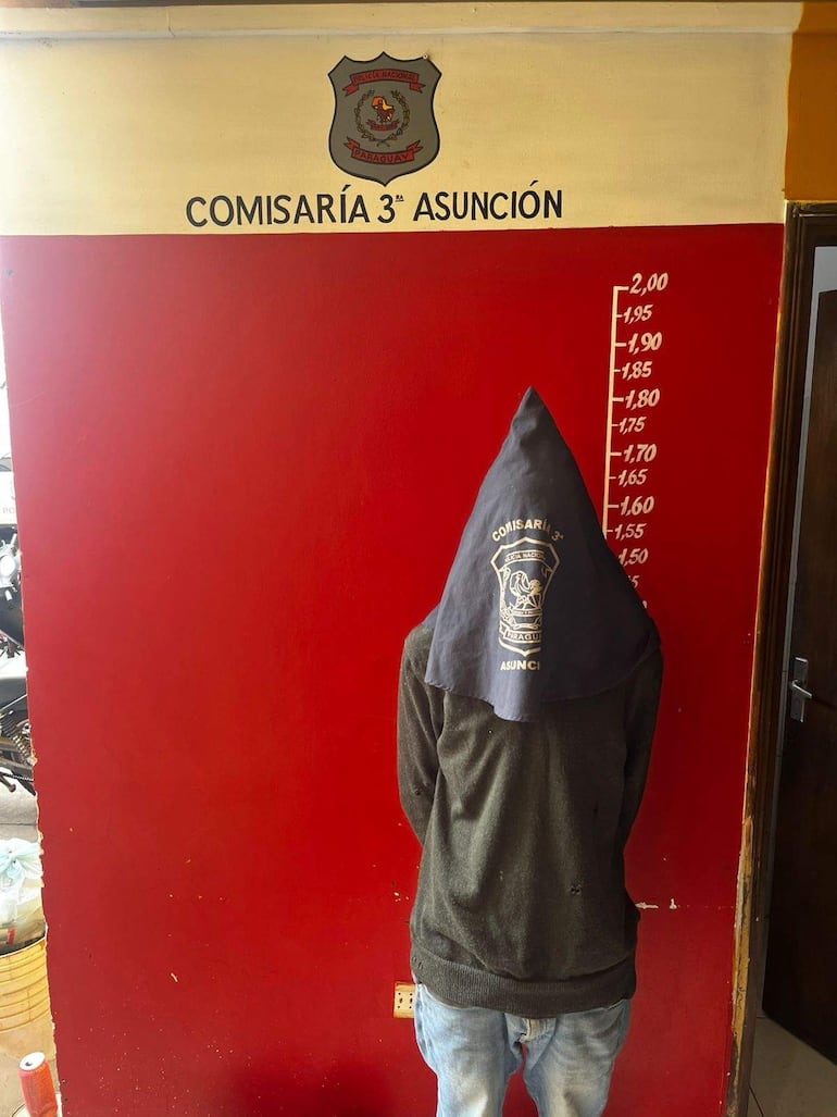 Hombre de espaldas con capucha oscura en la Comisaría 3ª de Asunción, con paredes rojas y amarillas y escudo policial.