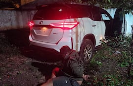 Hombre vestido con sudadera oscura y pantalones cortos, esposado sobre la hierba cerca de una SUV blanca con luces traseras encendidas.