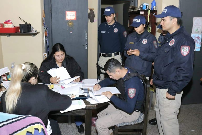 Grupo de policías en uniforme en una oficina desordenada, una mujer revisa documentos mientras otros observan con atención.