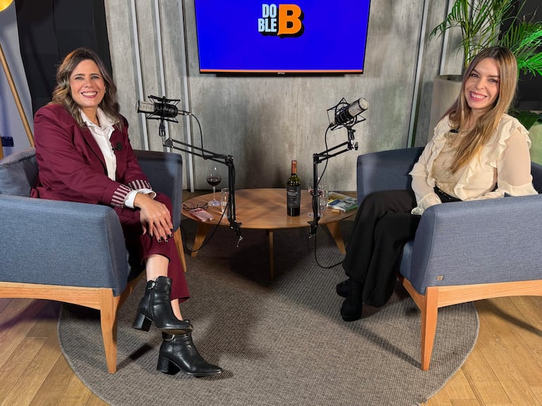 Kattya González, con traje burdeos, y Belén Bogado, en blusa blanca, sonríen sentadas en sillones azules en set de grabación.