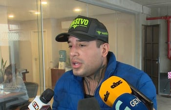 Hombre con chaqueta azul y gorro negro, tatuajes visibles, rodeado de micrófonos en oficina moderna, mientras habla con reporteros.