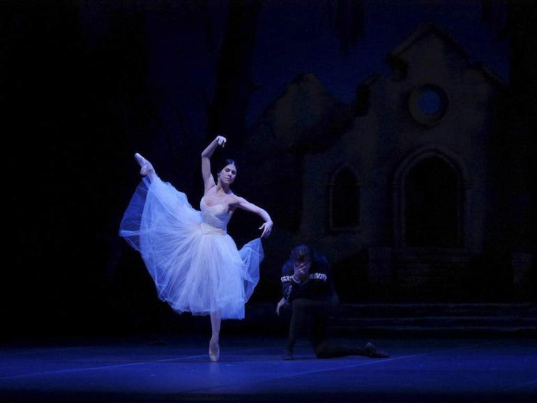 Bailarina en tutú blanco eleva su pierna, mientras un bailarín de rodillas expresa conexión emocional con ella en un escenario oscuro.