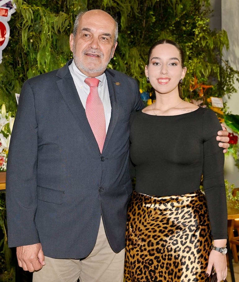 Hombre canoso en traje oscuro con corbata roja y mujer con vestido negro y falda de leopardo, sonríen frente a un fondo floral.