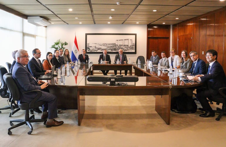 Trece personas en trajes formales alrededor de una mesa, con banderas y un mural en el fondo, en un ambiente colaborativo.