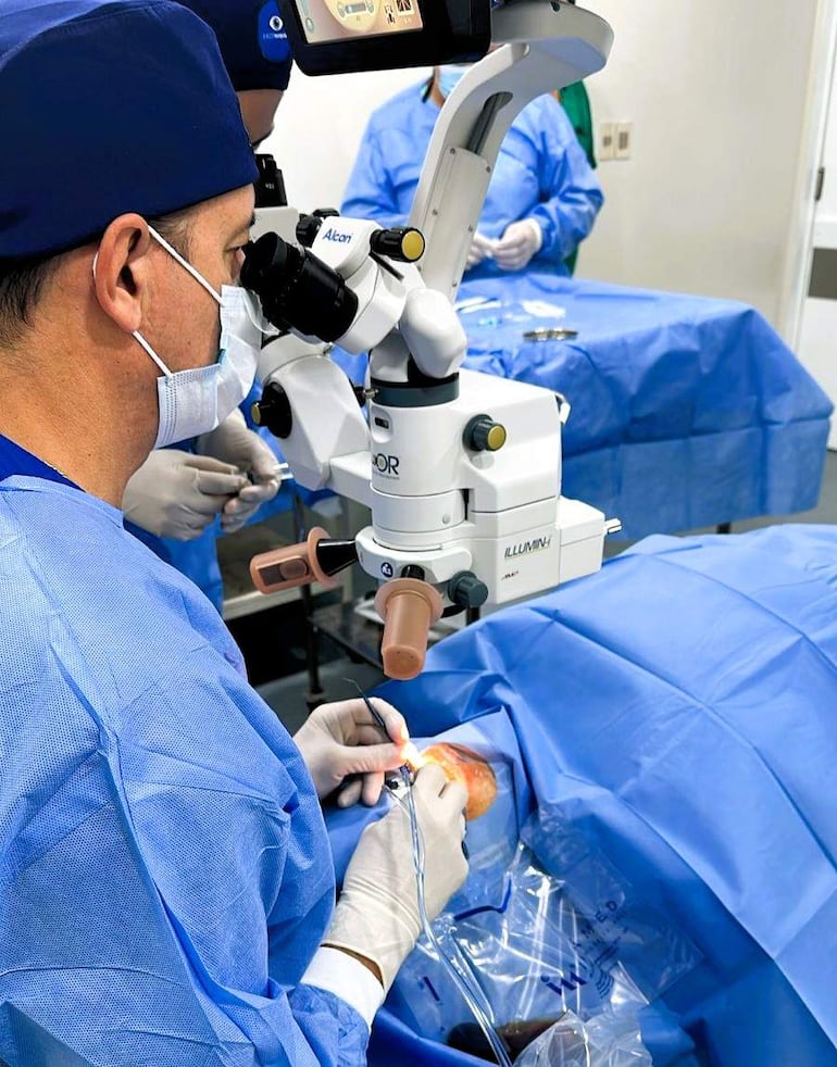 Médico de cabello corto con gorro azul, operando con microscopio en quirófano, acompañado de equipo de salud en vestimenta azul.